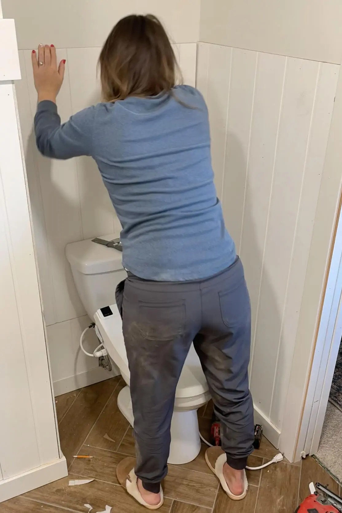 A woman installing vertical shiplap wall panels around a toilet in a bathroom remodel. She is pressing the panel into place while wearing casual DIY work clothes and cozy slippers. The wooden herringbone floor and unfinished trim show the progress of the renovation.